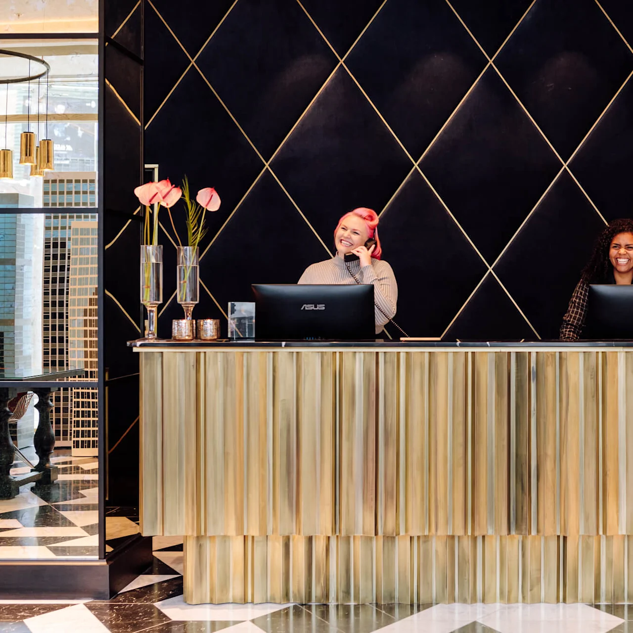 Reception desk with two smiling individuals; one talks on the phone. Behind, a geometric black and gold wall decorates the space. Pink flowers in glass vases are on the desk.