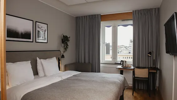 En pent oppredd seng med grått sengetøy står i et hotellrom. Til høyre står et skrivebord med en vannkoker og lampe, med et vindu som vender mot bytak.