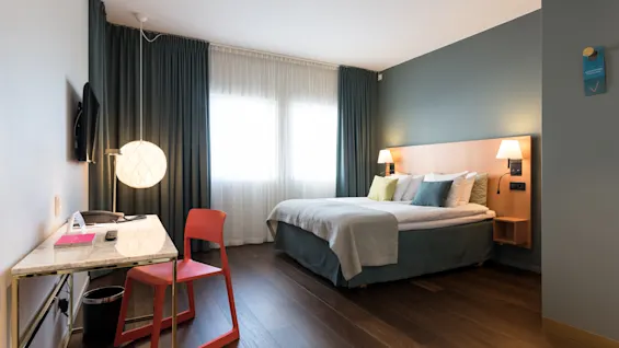 A neatly made bed stands against a green accent wall, with nightstands and lamps, in a modern, well-lit room with a desk, chair, and large window adorned with curtains.