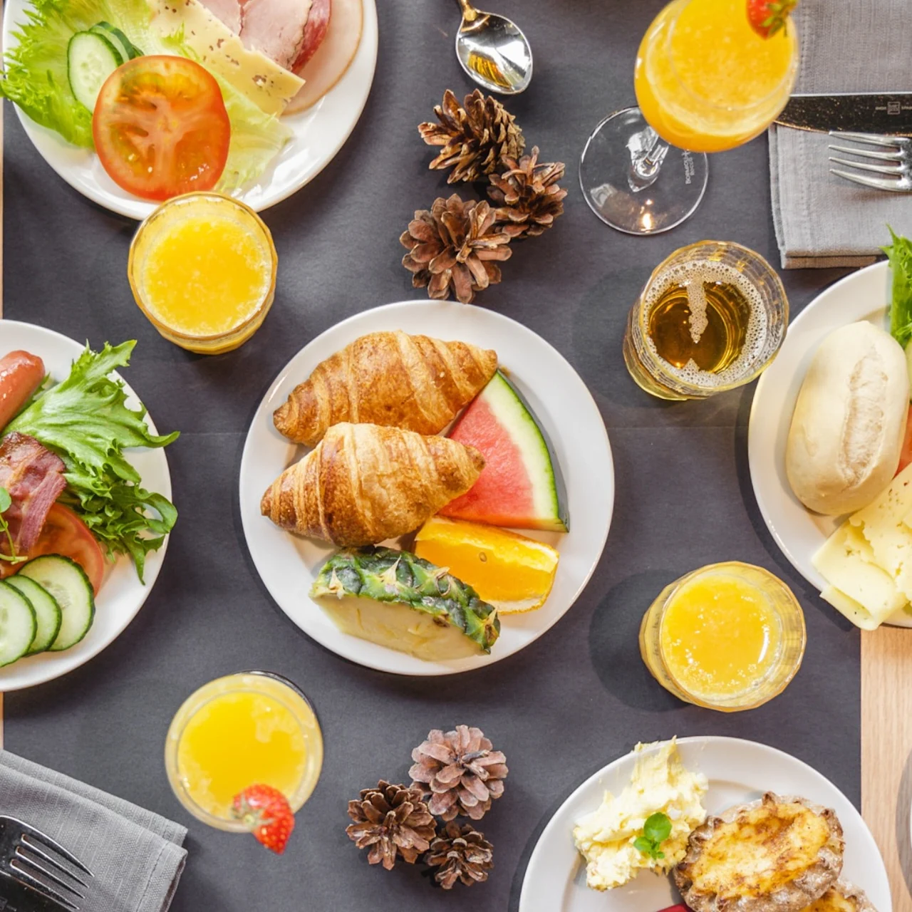 Plates of breakfast foods including croissants, sausages, and cheese arranged on a table with drinks and pinecones, surrounded by gray napkins and utensils.