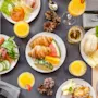 Lautasia aamiaista, mukaan lukien croissantteja, makkaroita ja juustoa, järjestetty pöydälle juomien ja käpyjen kanssa, harmaiden lautasliinojen ja ruokailuvälineiden ympäröimänä.