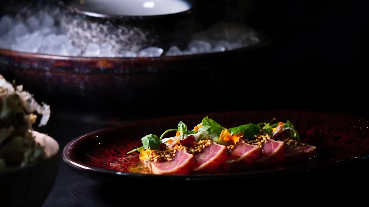 Skiver av brunet tunfisk er elegant lagt opp på en mørk tallerken, toppet med sesamfrø og garnert med friske urter og spiselige blomster, i et svakt opplyst matmiljø.
