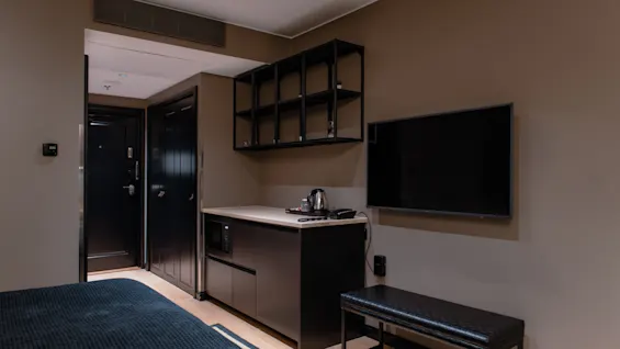 Hotellrom med koselig seng, praktisk kjøkkenkrok med mikrobølgeovn og vannkoker, stor TV og stilig benk.