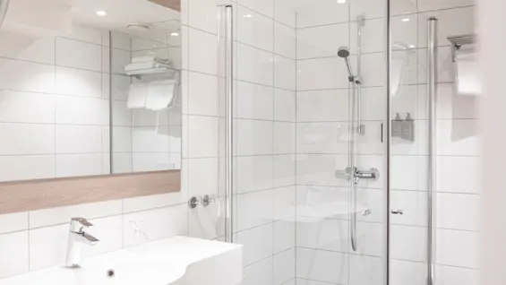 Et lyst, moderne hotellbad med hvit vask, stort speil og en dusj med glassvegger og friske håndklær.