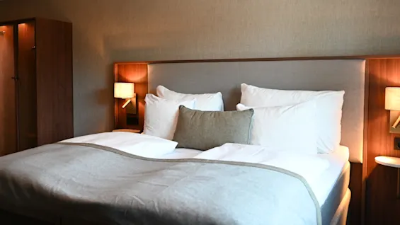 Et komfortabelt hotelværelse med en pænt redt seng, bløde puder og varm belysning fra sengelamper, der indbyder til afslapning.