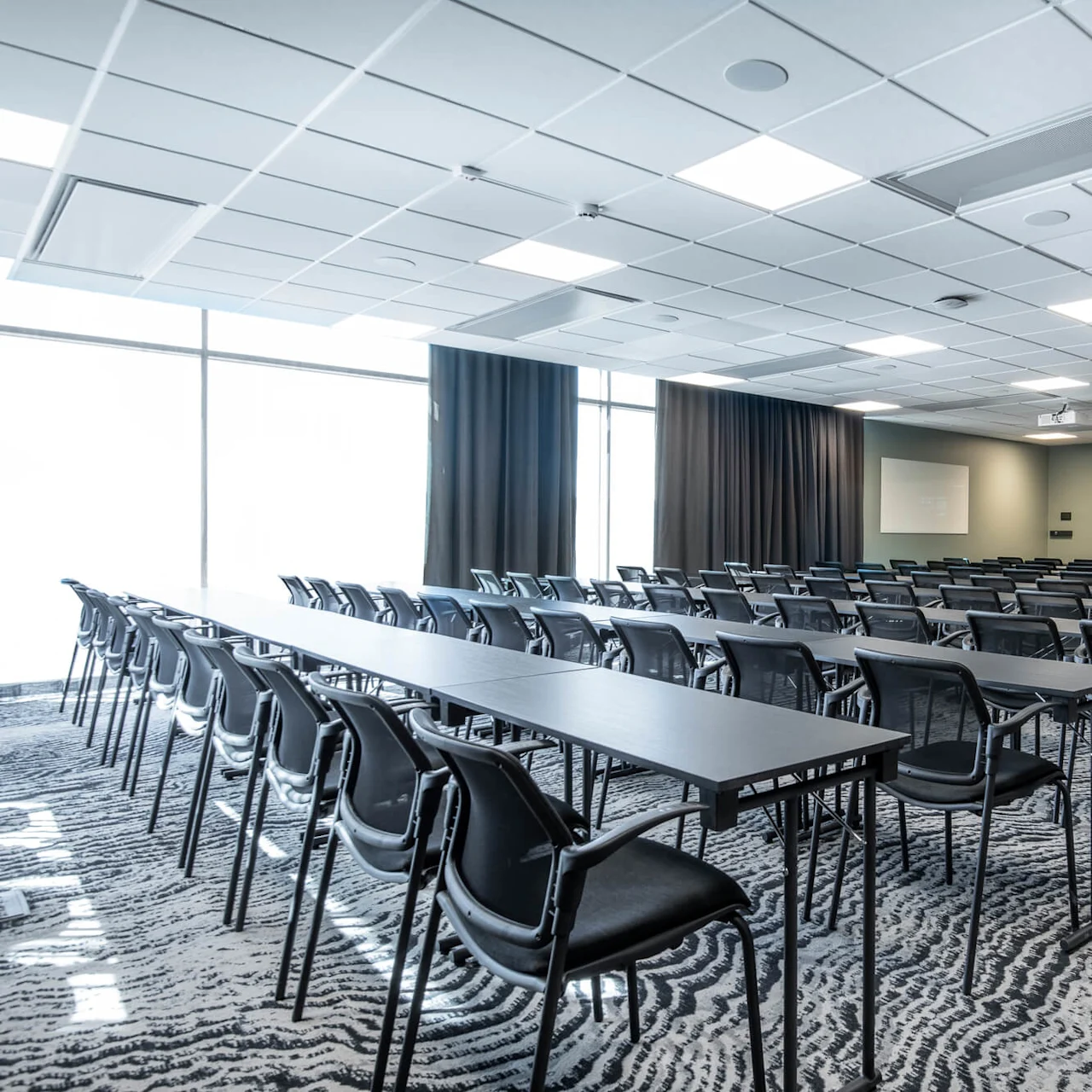 Rows of empty black chairs and tables face whiteboards and projectors in a spacious, modern conference room with patterned carpet and large windows.
