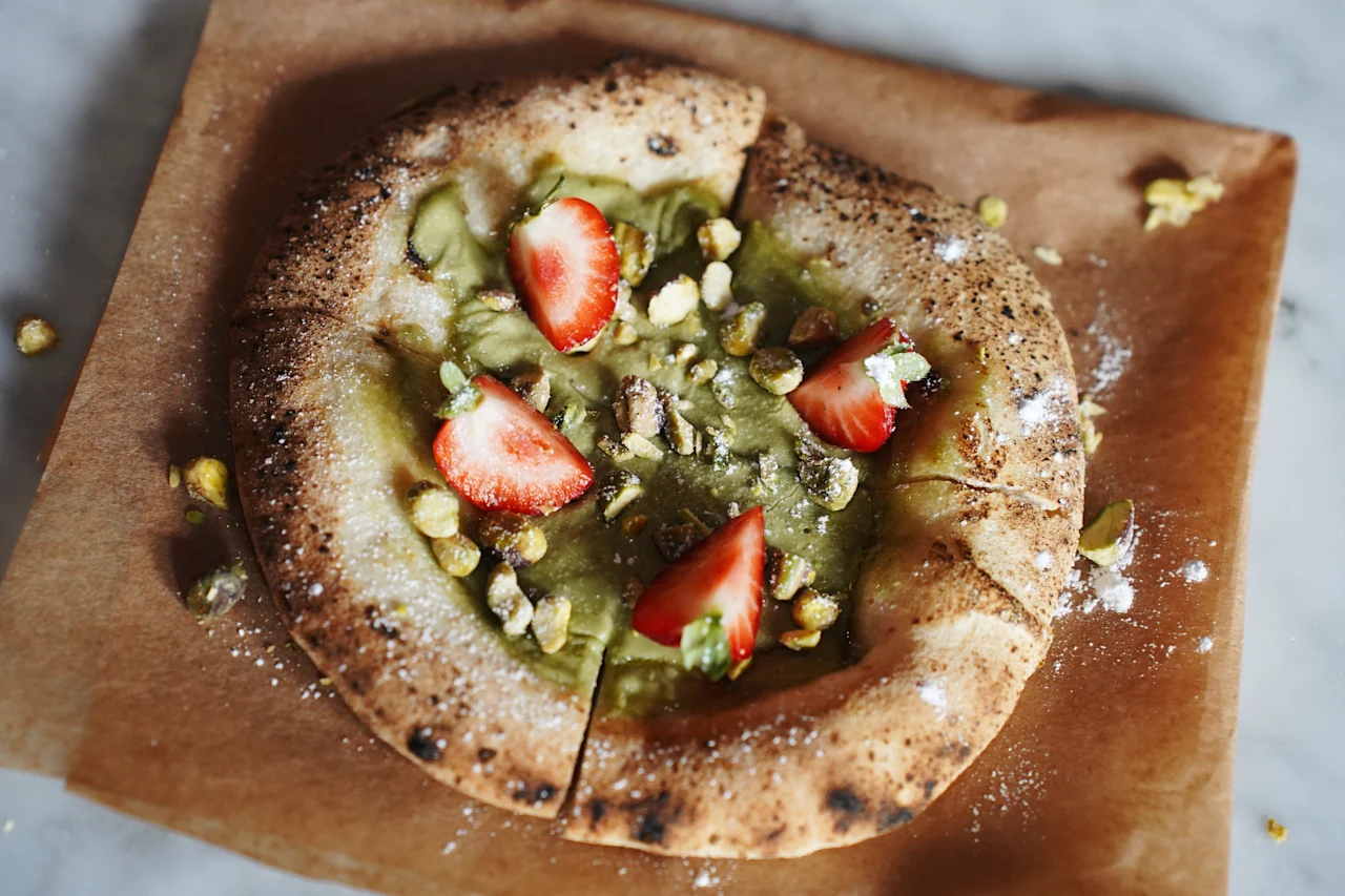 A round pastry topped with green spread, sliced strawberries, and chopped pistachios rests on brown parchment paper, placed on a marble surface.