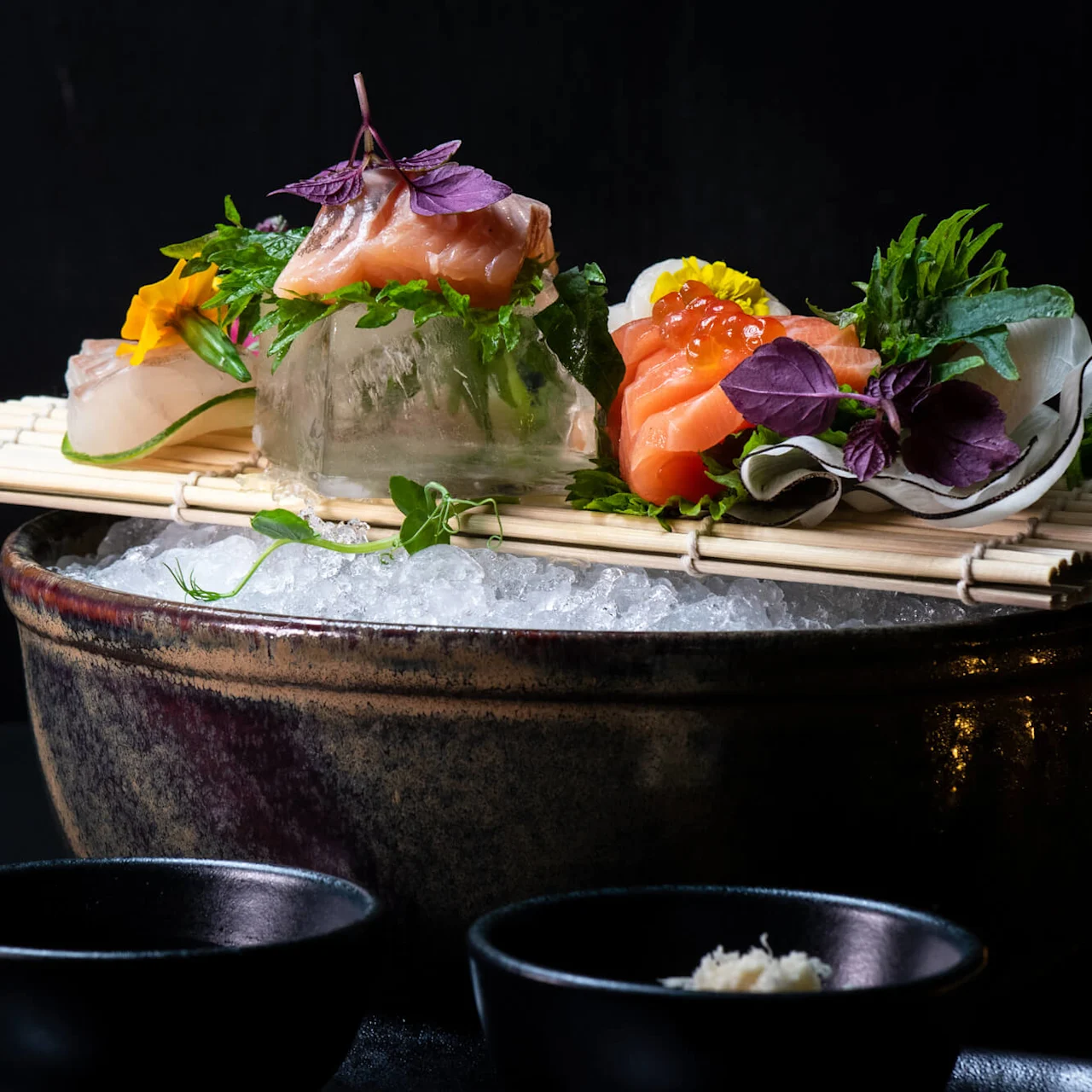 Sushi og sashimi vakkert arrangert på knust is med garnityr, plassert på en bambusmatte over en keramisk bolle i en mørk, minimalistisk setting. To små sorte boller er i forgrunnen.