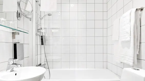 Et hvitt bad har en servant med et speil over, et badekar med dusjhode og håndklær som henger på en stang. Rommet har flislagte vegger og glasshyller.