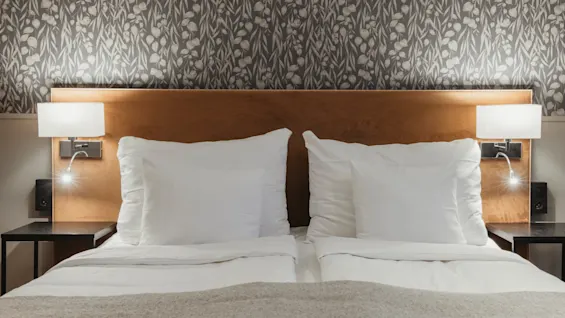 En bekväm hotellsäng med vita lakan, en sänggavel i trä och väggmonterade lampor, mot en mönstrad tapet.