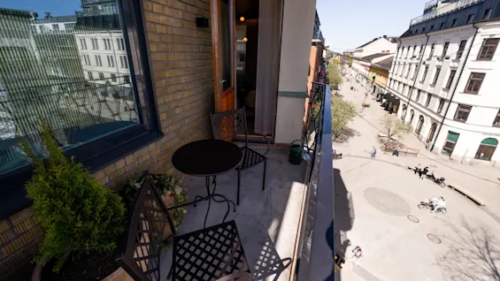A small balcony features a round black table with two metal chairs. It overlooks a lively street with people walking and cycling past historic buildings. 