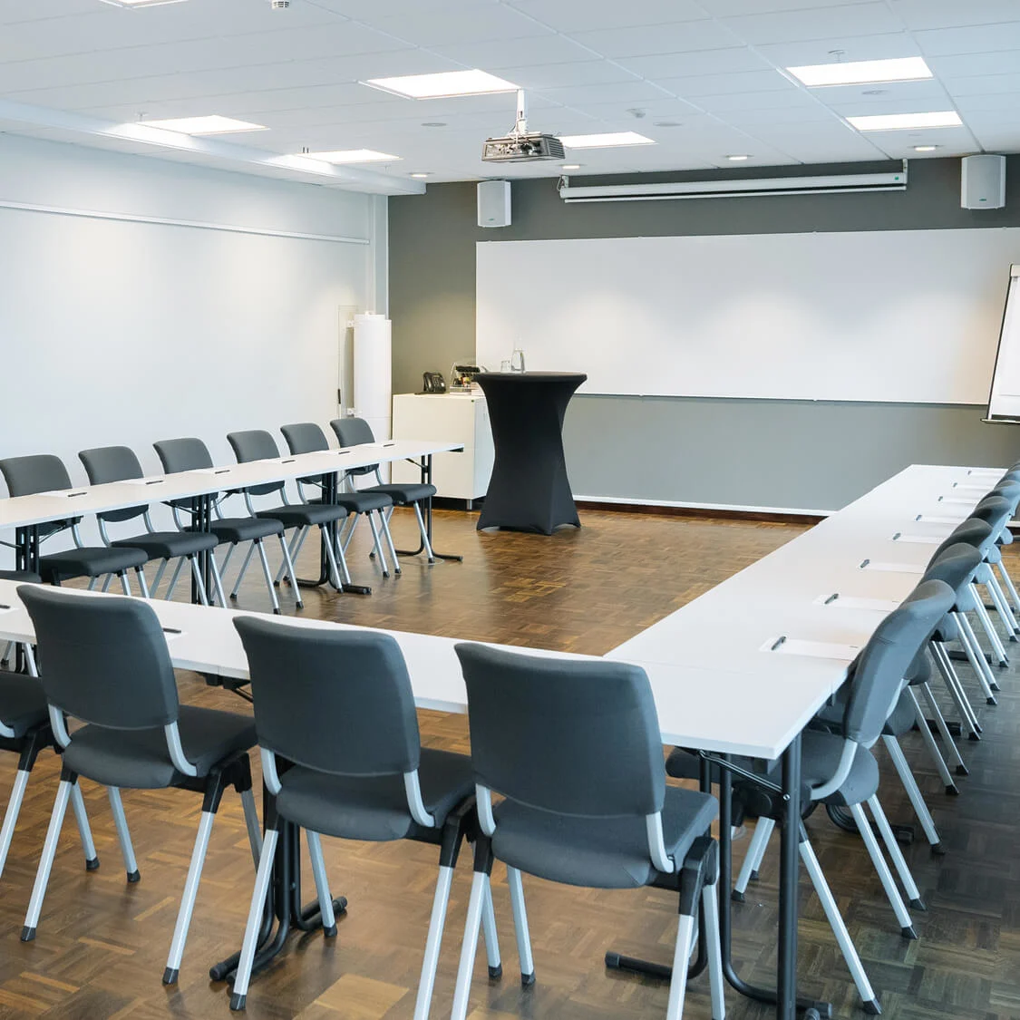 Mötesrum med U-formade bord och grå stolar, vända mot en stor whiteboard. En dörr med nödutgångsskylt finns till vänster. Ett blädderblock står i hörnet.