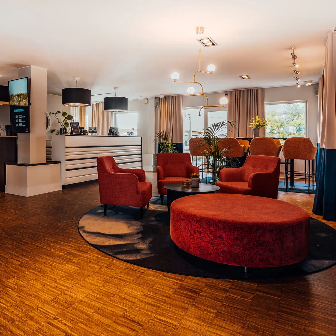 Ett modernt lobbyområde med röda fåtöljer, runda bord, trägolv och stämningsbelysning.
