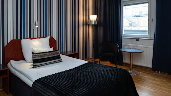 En pænt redt enkeltseng med stribet sengelinned står op ad en stribet væg. Et vindue med mørke gardiner, en lampe og et lille bord med en stol er i nærheden.