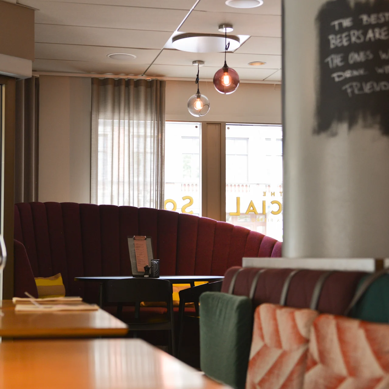 En restaurantinteriør med hyggelige siddepladser, vinglas på borde, hængelamper og tavletekst: ”THE BEST BEERS ARE THE ONES WE DRINK WITH FRIENDS,” skaber en varm atmosfære.