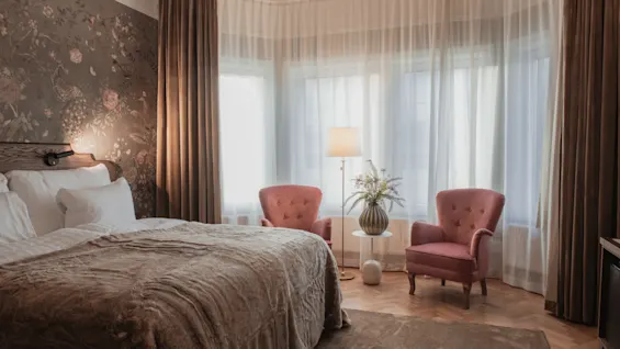 Home Hotel Baltzar - Deluxe Double. A cozy hotel room with a large bed, floral wallpaper, and two pink armchairs by a window.