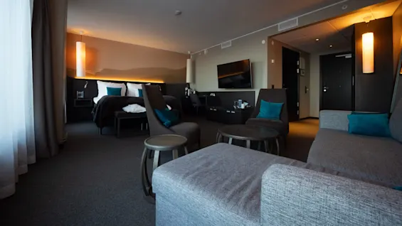 Et hotelværelse har en stor seng med turkise puder og et siddeområde med stole og sofa. Blød belysning og moderne interiør skaber en hyggelig stemning.