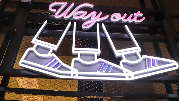 Neon signs displaying "ELEVATORS" in an arrow, above "Way out" and shoes graphic, mounted on a mesh background. Partial text "ROOMS" visible above.