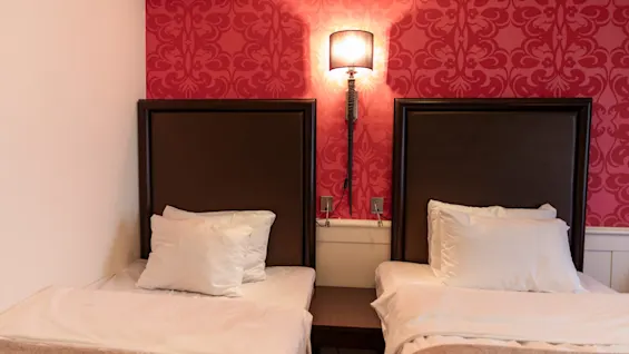 Two single beds with white bedding and dark headboards in a room with red patterned wallpaper and a wall lamp. Home Hotel Grand Bodø.