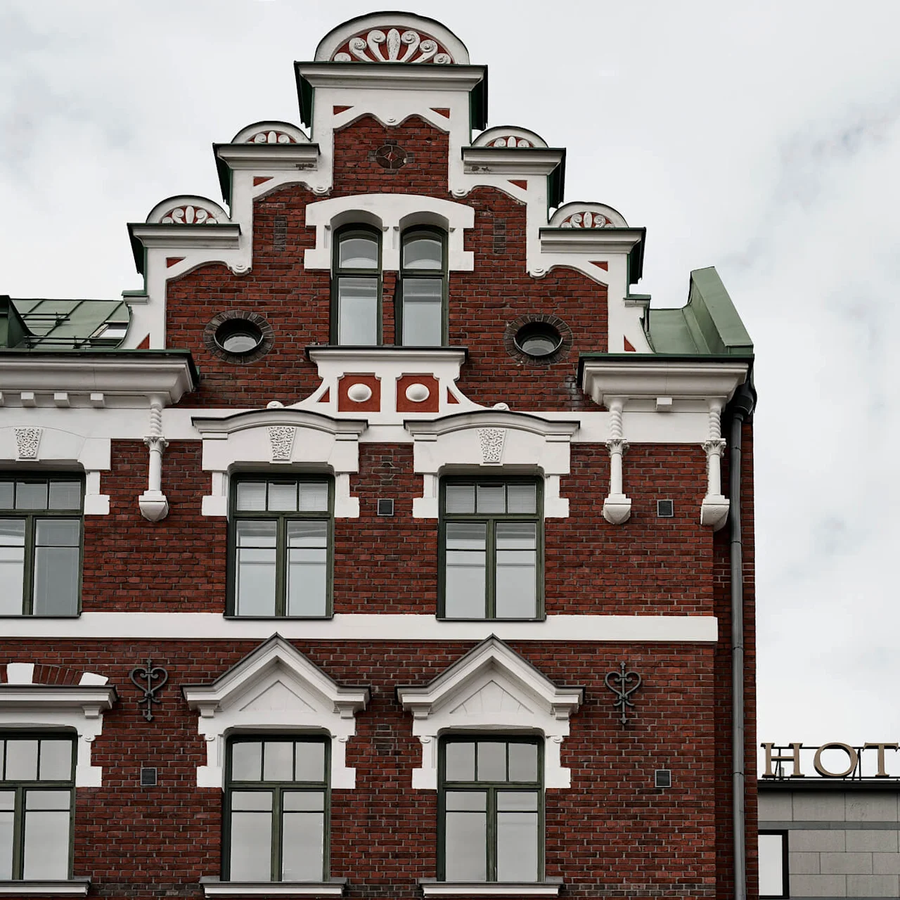 A historic red-brick building displays intricate white decorative accents and modern windows. Adjacent is a modern structure with a sign reading "HOTEL" under a cloudy sky.
