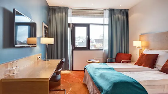 En seng med turkis teppe og oransje puter står i et hotellrom. Overfor er det et skrivebord med stol og flatskjerm-TV. Stort vindu med gardiner gir naturlig lys.