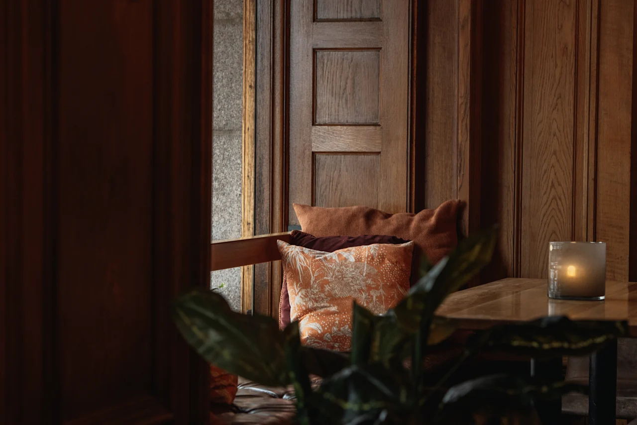 Ett mysigt, varmt upplyst hörn med träväggar, mjuka kuddar och ett glödande ljus på ett bord. Home Hotel Grand, Helsingborg.