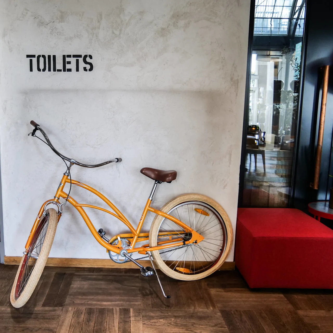 An orange bicycle rests against a wall inside a building, next to a red bench. The word "TOILETS" is stenciled on the wall, near a glass door.