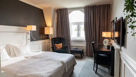 Et hyggeligt hotelværelse med komfortabel seng, lænestol og skrivebordsområde, badet i naturligt lys fra stort vindue.