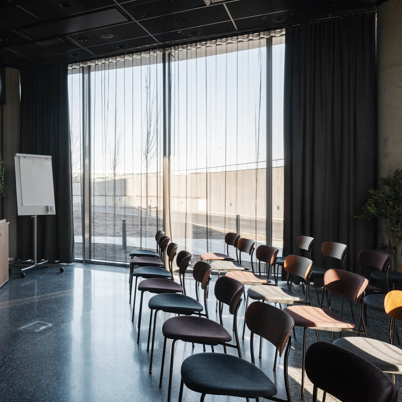 A conference room features rows of empty chairs facing a large screen. A flip chart stands nearby, with floor-to-ceiling windows allowing natural light inside.