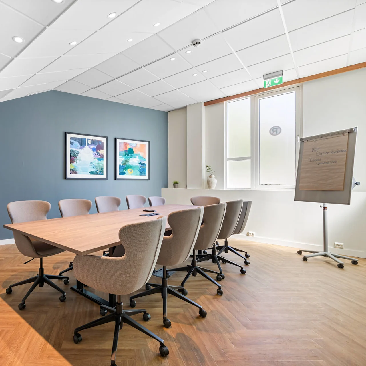 A modern conference room with a long table, chairs, and a flipchart. Natural light and artwork on the wall create a bright atmosphere.