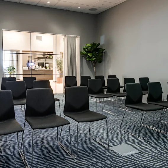 Rows of black chairs face an open doorway that leads to a bright room with plants. A wall-mounted clock hangs beside a closed door, creating a conference setting.