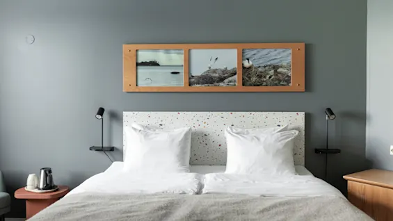 A cozy hotel room with a bed, a speckled headboard, and a wooden frame above it displaying three coastal landscape pictures.