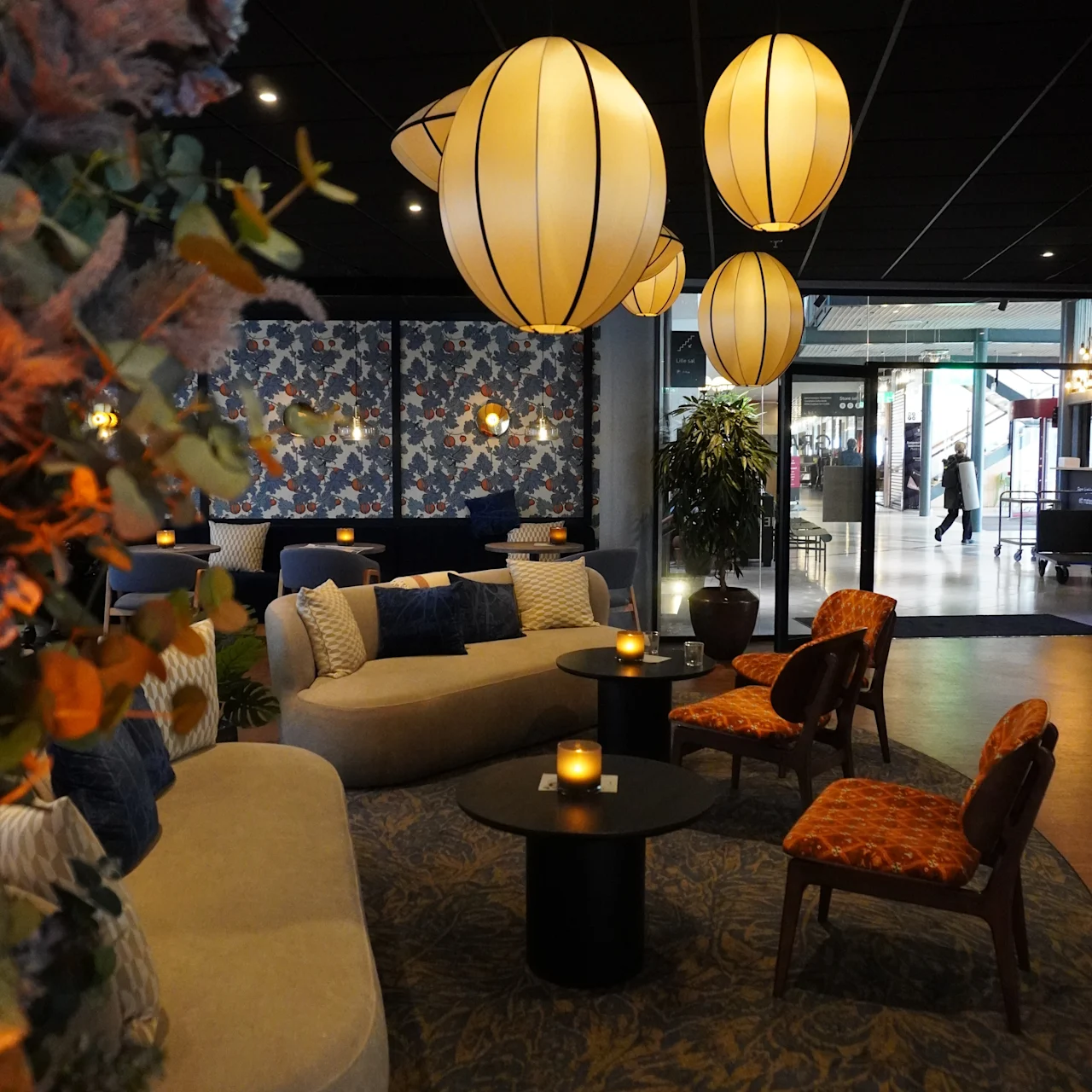 Taklampor lyser upp en mysig lounge med soffor, stolar och bord. Blommiga tapeter och växter förstärker den inbjudande atmosfären. En glasentré visar förbipasserande.
