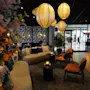 Taklampor lyser upp en mysig lounge med soffor, stolar och bord. Blommiga tapeter och växter förstärker den inbjudande atmosfären. En glasentré visar förbipasserande.