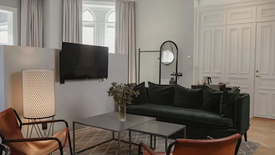 En modern vardagsrum har en grön soffa med mörka kuddar, en väggmonterad TV och två bruna stolar runt ett bord med en vas. Mjuk belysning och neutral inredning skapar en mysig atmosfär.