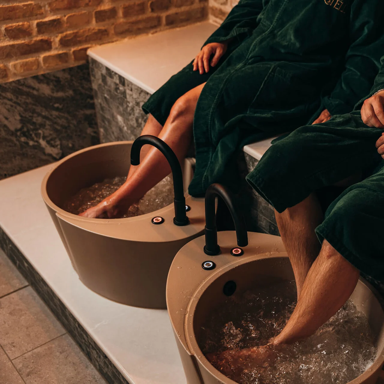 Two people in green robes soak their feet in bubbling foot baths, seated on a tiled bench in a spa setting. "FRIÉL" is embroidered on their robes.