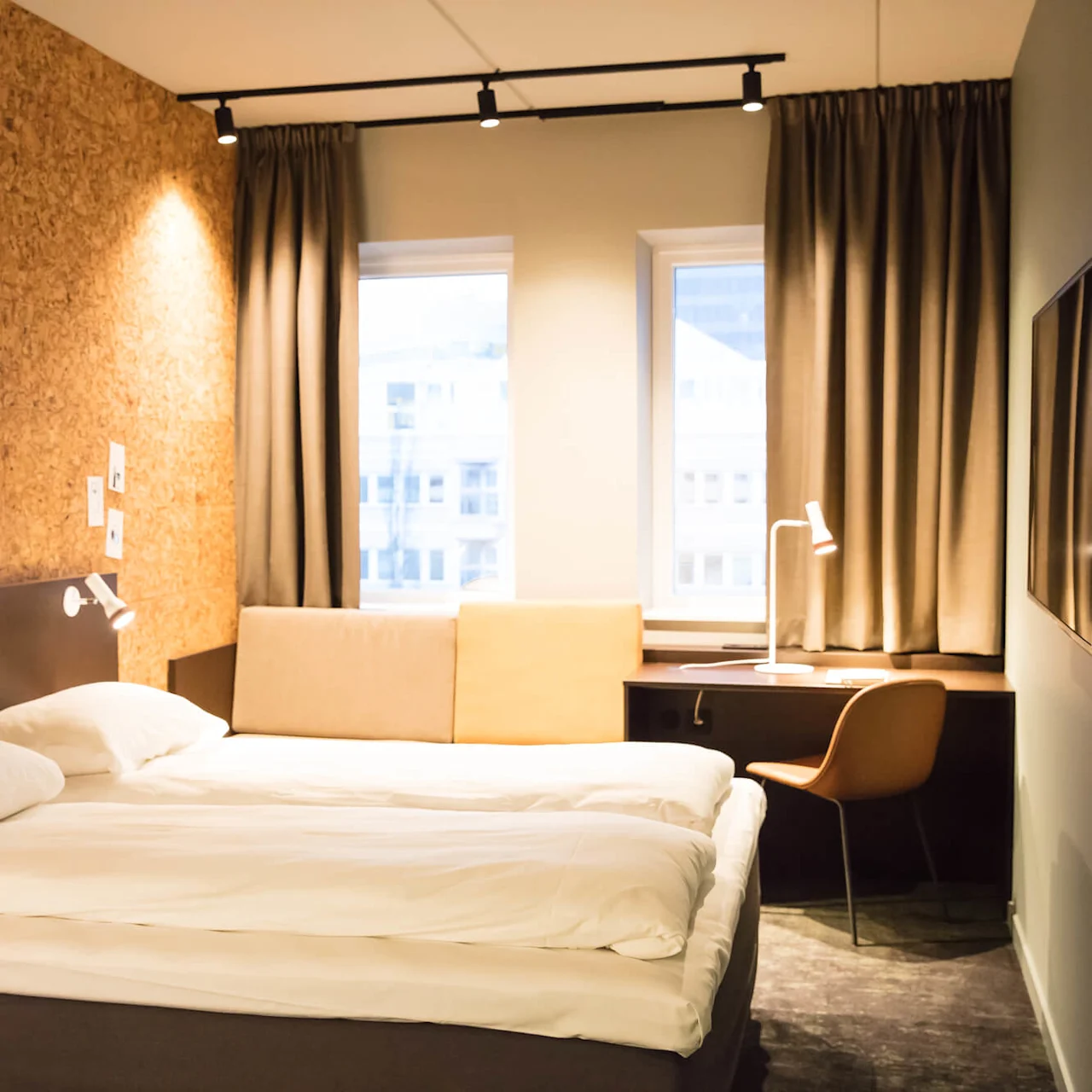 A double bed is positioned against a cork-textured wall, with bedside lamps. Adjacent, a desk with a chair and lamp sits by two curtained windows, in a minimalist hotel room.