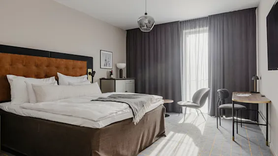En pænt redt seng med hvidt sengetøj i et moderne, minimalistisk rum med skrivebord, stole og et stort vindue med mørke gardiner. Et indrammet billede hænger over et skab.