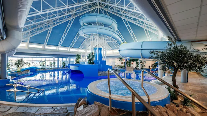Indoor water park with a winding blue slide above a large pool. Surrounding the area are plants and stone pathways under a high, triangular glass ceiling.