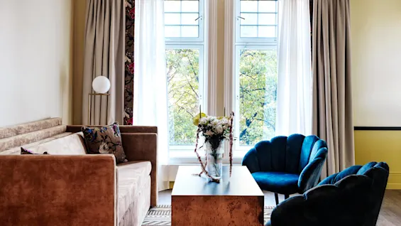 A brown sofa faces a wooden table with a flower arrangement, flanked by blue chairs. Behind, large windows with curtains reveal green trees outside.