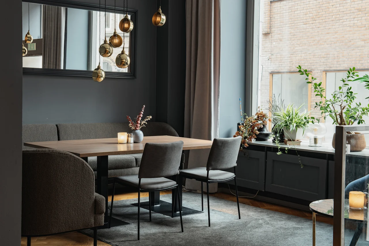 Et moderne spiseområde med et langt trebord, komfortable sitteplasser og varm belysning, med planter ved et stort vindu.