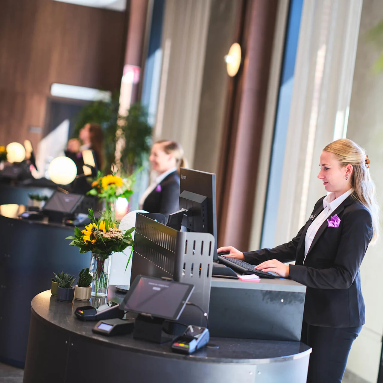 Receptionister ved buede borde arbejder ved computere, omgivet af dekorative planter og blomster, i en lyst oplyst lobby.