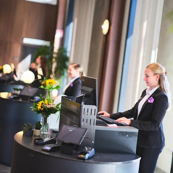 Receptionister vid böjda skrivbord arbetar vid datorer, omgivna av dekorativa växter och blommor, i en ljust upplyst lobby.