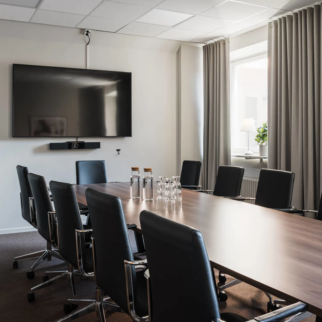 Ett långt konferensbord med svarta stolar står mot en stor väggmonterad skärm. Vattenflaskor och glas är placerade på bordet. Rummet pryds av en krukväxt och gardiner.