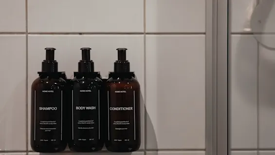 Tre mörka flaskor med schampo, kroppstvål och balsam monterade på en kaklad duschvägg. Etiketterna lyder 100% Vegan.