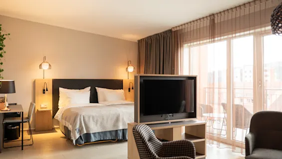 Ett rymligt hotellrum med en säng och kuddar till vänster, en TV som vetter mot en sittgrupp i mitten och en balkong utanför en glasdörr till höger.