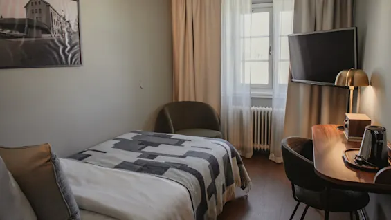 Ett mysigt hotellrum med en enkelsäng, en skrivbordshörna och en väggmonterad TV, som erbjuder en bekväm vistelse.