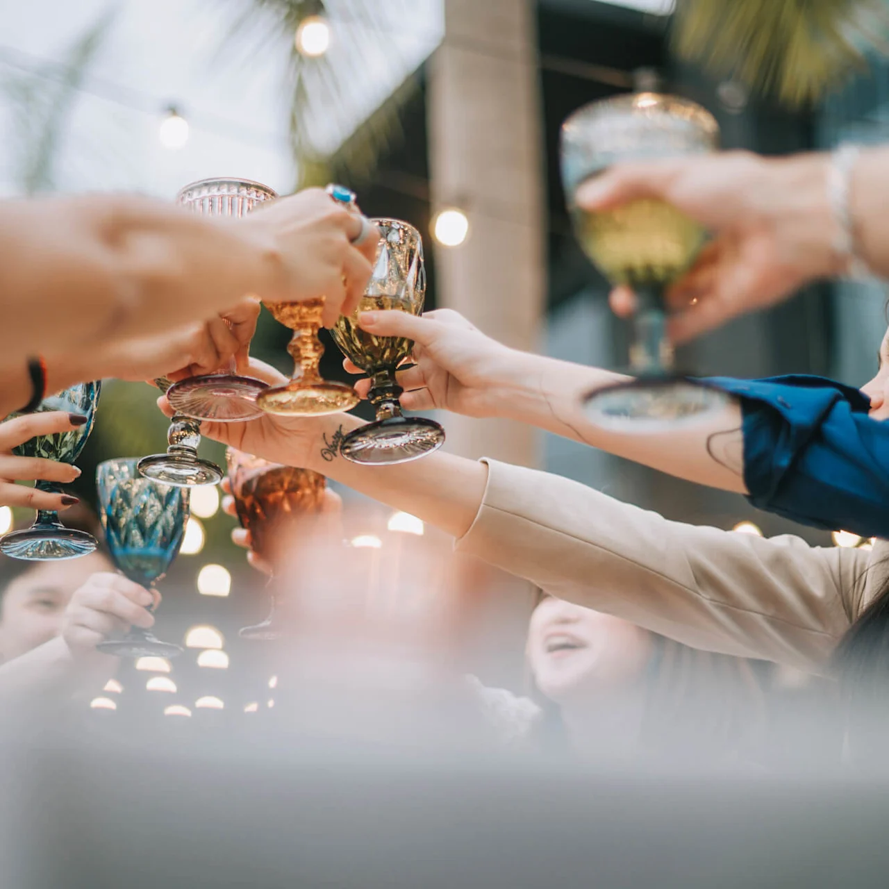 Hands raising colorful glasses in a toast, with smiling people and string lights in the background setting a festive outdoor gathering ambiance.