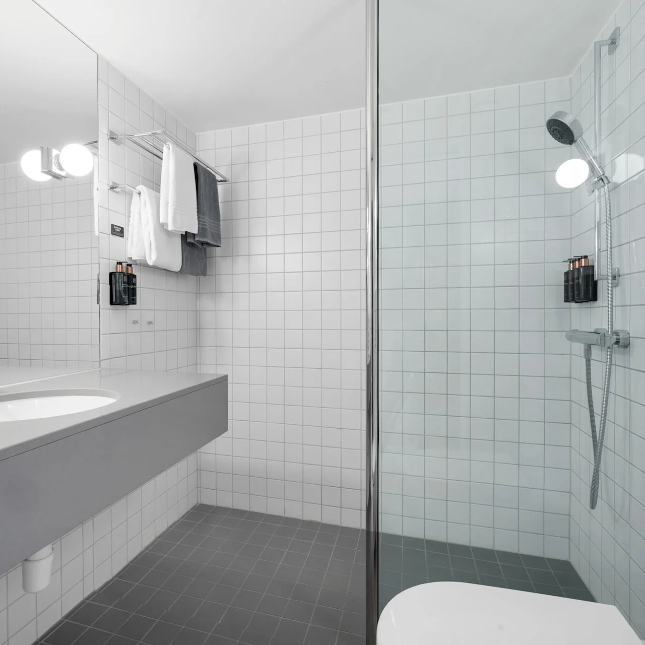 Et bad med en moderne vask og stort speil. I nærheten ses en dusj med glassvegger og krom dusjhode. Håndklær og toalettartikler er pent plassert på hvite flisvegger.