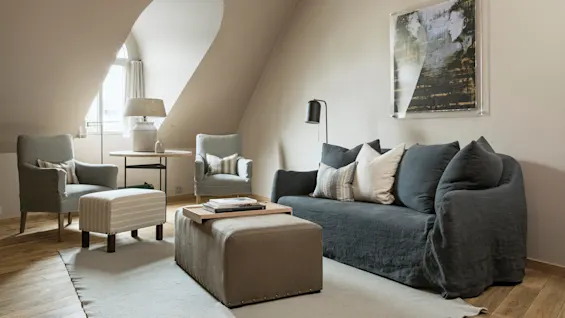 A cozy living room features a dark sofa with pillows, two armchairs, and a small table by a window. A framed artwork hangs on a sloped beige wall.